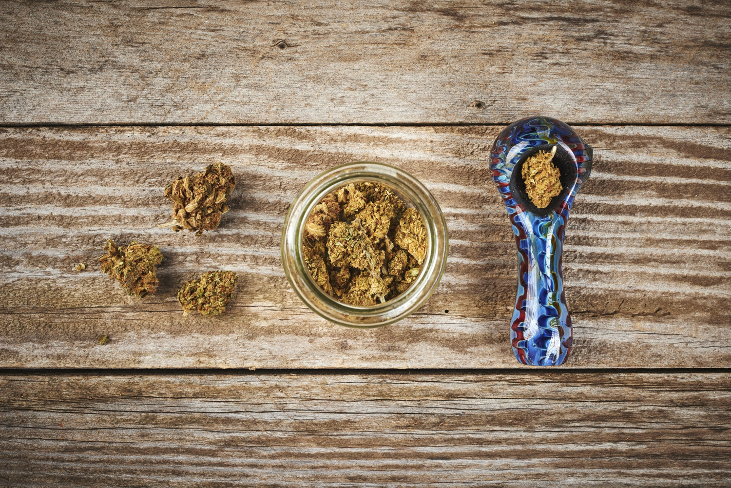 A glass pipe sits on a wood table beside a jar of fresh cannabis buds