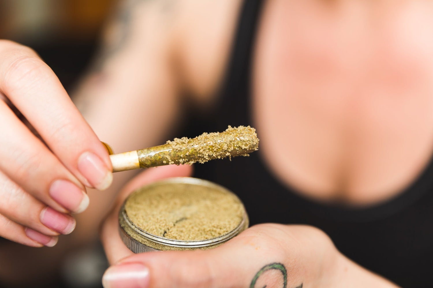 A woman rolls a kief joint. A grinder full of cannabis trichomes is in her left hand while a kief joint is in her right.