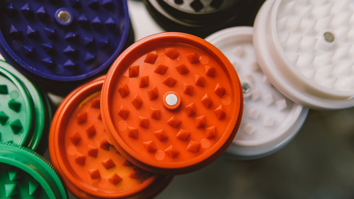 Clean plastic weed grinders sit in a pile. One is orange, one is white, one is purple, one is green and one is black. 