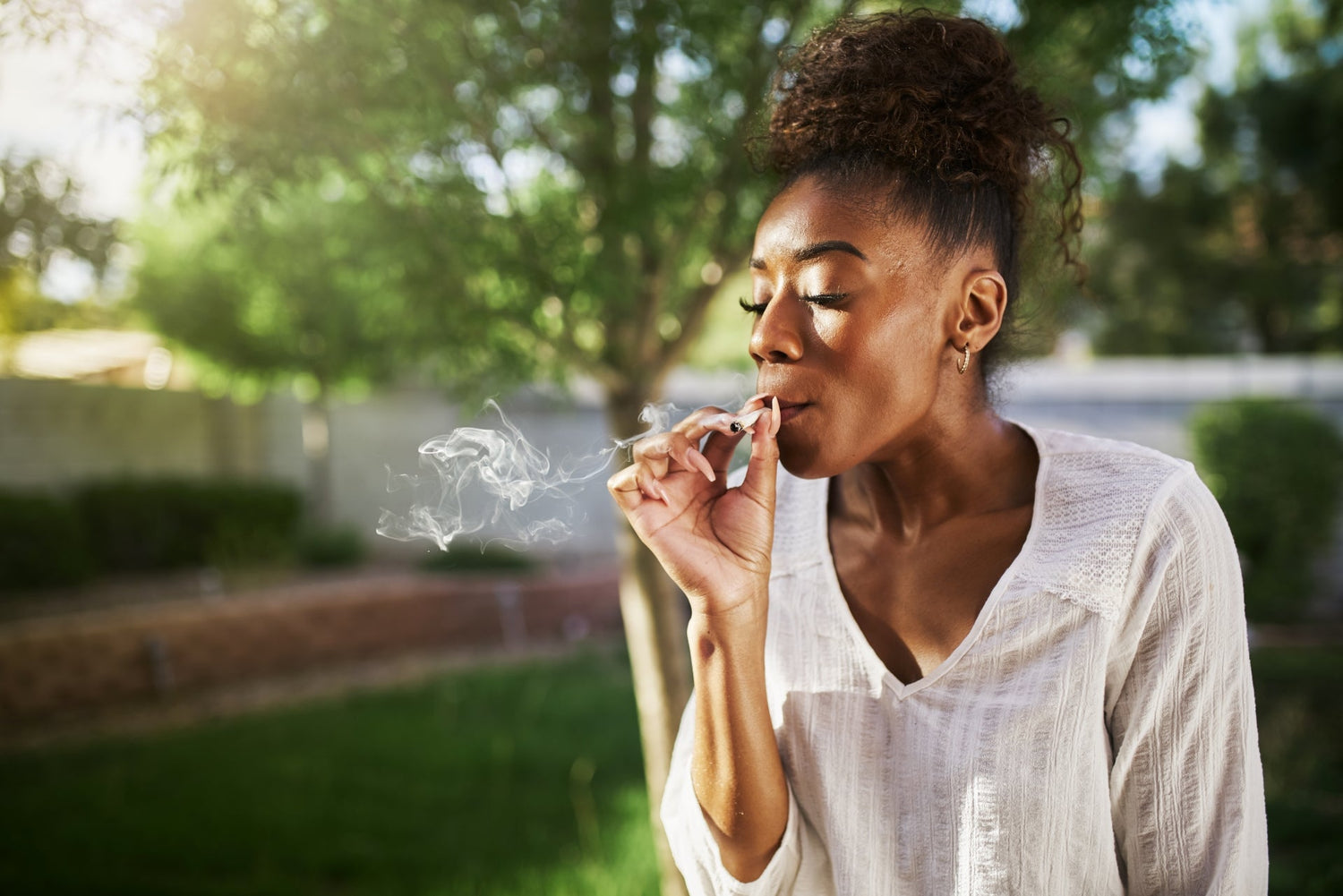 A black woman smokes a joint without a filter at a park on a summer's day