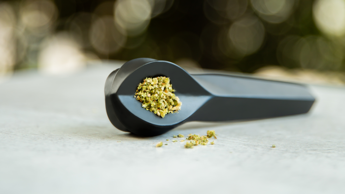 A pipe is packed with ground cannabis. It is sitting on a cement bench against a background of greenery