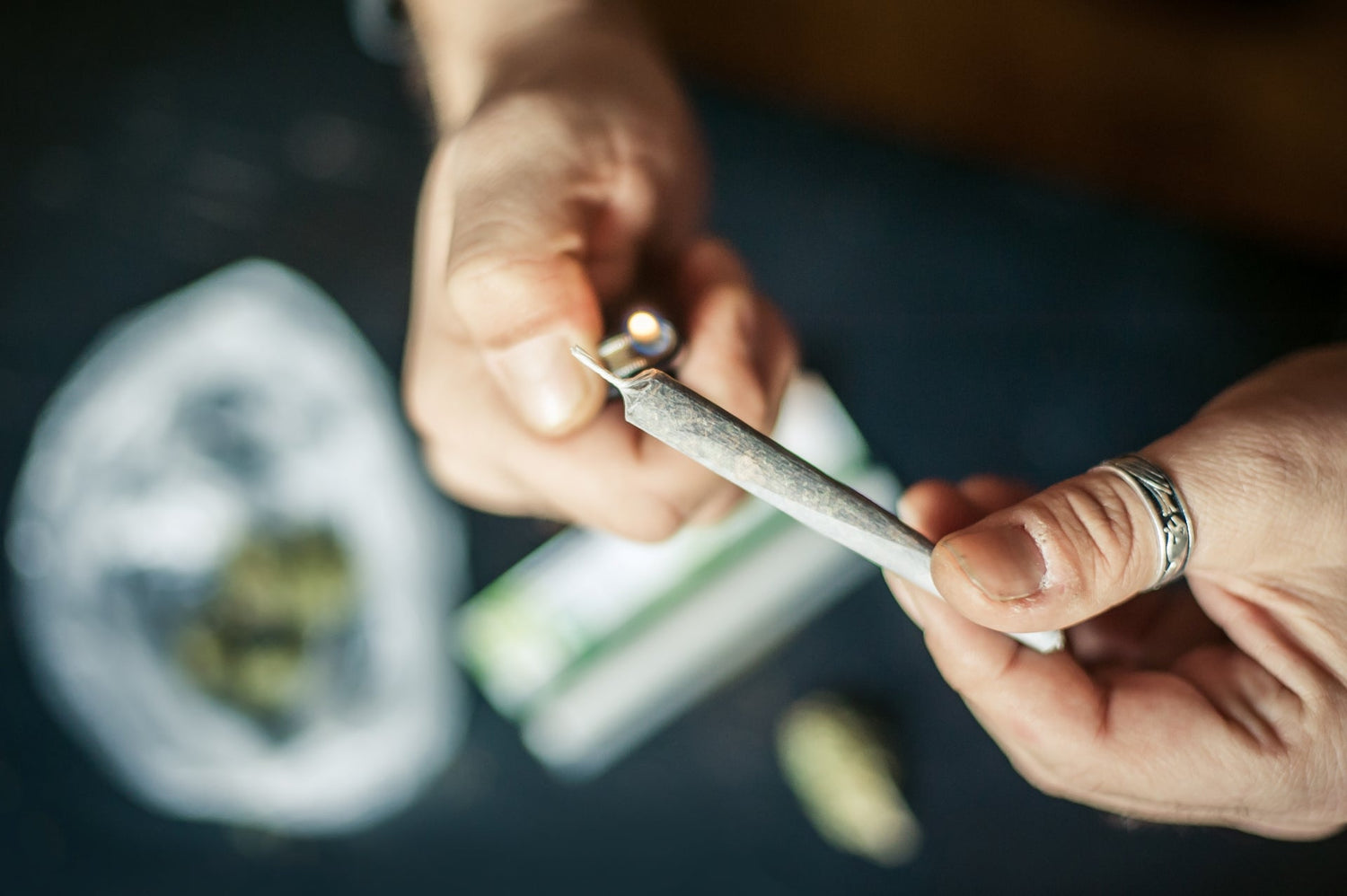 A man's hands hold a perfectly rolled thin joint. He has a ring on his left thumb and is using his right hand to light the joint.
