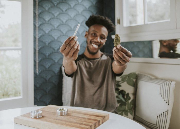 A smiling handsome black man holds a cone joint and cannabis nug to the camera. He sits in a kitchen and is about to roll another cone joint