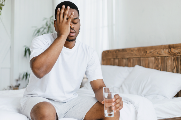 black man waking up in bed feeling hangover holding a glass of water